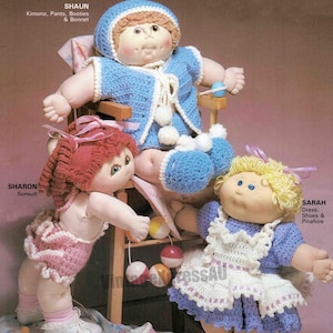 May include: Three Cabbage Patch Kids dolls wearing crocheted outfits. The doll on the left is wearing a pink sunsuit, the doll in the middle is wearing a blue kimono, pants, booties, and a bonnet, and the doll on the right is wearing a purple dress, white pinafore, and purple shoes.