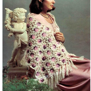 Könnte beinhalten: Ein cremefarbener gehäkelter Schal mit einem Blumenmuster in Rosa und Lila. Der Schal hat einen Fransenrand und ist über einer Person drapiert, die ein rosa Kleid trägt. Im Hintergrund befindet sich eine Statue eines Engels.