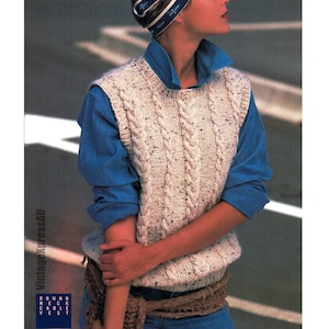 May include: A white cable knit waistcoat with a round neckline. The waistcoat is worn over a blue button-down shirt and a brown belt.