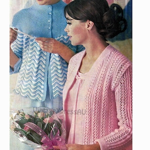 Op de afbeelding: Een vintage afbeelding met twee vrouwen in gebreide vesten. De ene vrouw draagt een lichtblauw vest met een geschulpte rand, de andere een roze vest met open voorkant. De afbeelding heeft een retro-esthetiek, met een boeket bloemen op de voorgrond. Vintage Xpress AU.