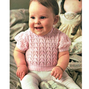 Può includere: Una bambina che indossa un maglione rosa lavorato a maglia con un motivo a pizzo. Il maglione ha maniche corte a sbuffo e un collo rotondo. La bambina è seduta su una coperta con un motivo floreale.