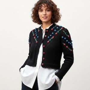 May include: A black knit cardigan featuring a button-down front with bright red buttons. The shoulders and upper sleeves are decorated with embroidered green stems, small blue pom-poms, and red and white spotted mushroom-shaped embellishments.
