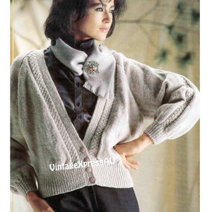 May include: A light grey knitted cardigan with a V-neck and button fastening. The cardigan features a cable knit pattern and a textured design. The model is wearing a white scarf with a decorative pin.