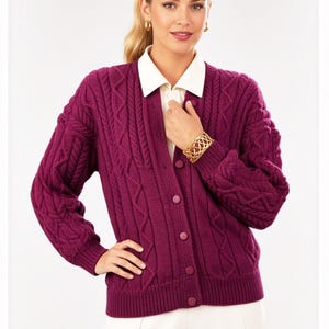 May include: A deep magenta cable-knit cardigan featuring a V-neckline, long sleeves with ribbed cuffs, and a front button closure. The knit pattern consists of vertical braided cables and textured panels. The garment is worn over a white collared shirt.
