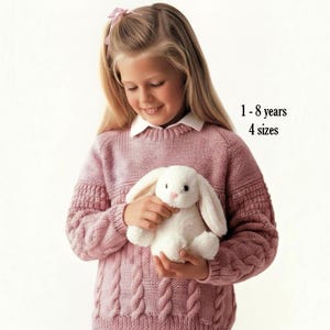 May include: A dusty rose long-sleeved knit sweater featuring a cable knit pattern on the lower section and a smooth knit upper section. The sweater includes the text 1 - 8 years 4 sizes. A white plush rabbit toy is held against the front of the sweater.