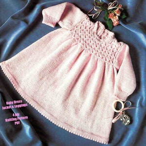 May include: A pink knitted baby dress with a lacy pattern on the bodice and a scalloped hem. The dress is laid out on a blue fabric background. The text "Baby Dress Jacket & Leggings 4 ply Knitting Pattern PDF" is printed below the dress.
