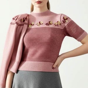 May include: A short-sleeved pink knit sweater featuring a ribbed crew neckline, horizontal dark pink stripes across the torso, and a floral embroidery pattern along the upper chest. A matching pink knit cardigan is draped over one shoulder.