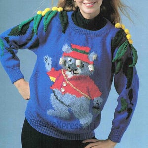 Puede incluir: Un suéter de punto azul con un koala que lleva un sombrero rojo y una banda. El suéter tiene hojas verdes y amarillas en los hombros.