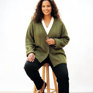 May include: An olive green cable-knit cardigan featuring a V-neckline, a single large button closure at the center, and long sleeves with folded cuffs. The garment is worn over a plain white V-neck top and paired with solid black trousers.