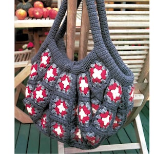 May include: A handmade, dark gray crochet tote bag with a floral pattern in red and white. The bag features a square granny stitch design and sturdy handles, perfect for carrying items. The bag is displayed outdoors.