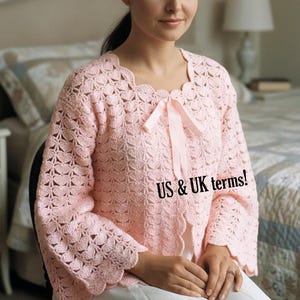 Pode incluir: Um cardigã de crochê rosa claro com borda festonada, mangas compridas e um laço de fita de cetim no decote. A peça apresenta um padrão de pontos em forma de leque repetido. O texto US & UK terms! é visível na imagem.