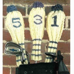 May include: Three cream-colored knitted golf club head covers with blue accents and pom-poms. Each cover has a different number: 1, 3, and 5. The golf clubs are in a black golf bag, set against a brick wall.