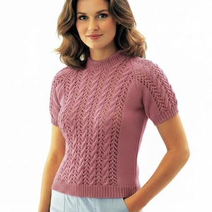May include: A short-sleeved, mauve-colored knit sweater featuring a mock neck, vertical cable knit patterns, and openwork lace detailing. The garment has ribbed trim at the neckline, sleeve cuffs, and hem, with solid knit side panels.