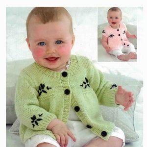 May include: A baby wearing a light green knitted cardigan with black floral embroidery and black buttons. The cardigan is paired with white shorts. A smaller image shows a baby in a pink knitted top with similar floral embroidery and black buttons.