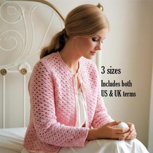 May include: A light pink long-sleeved crochet cardigan with a round neckline and a thin ribbon tie closure at the front. The text on the image reads: 3 sizes, Includes both US & UK terms. The garment features a repeating openwork stitch pattern.