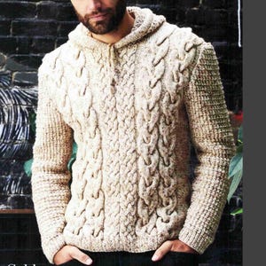 May include: A cream-coloured cable-knit hooded jumper with a zip. The jumper has a textured pattern of thick, interwoven cables and a ribbed knit design on the sleeves. The person is wearing black trousers.