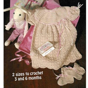Puede incluir: Vestido y patucos de bebé de ganchillo en color beige claro, con cintas rosas. Un conejito de peluche con peto rosa y una caja de regalo también están en la imagen. El texto dice "2 tallas para tejer a ganchillo 3 y 6 meses."