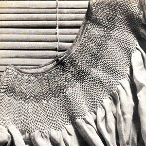 May include: A close-up of a white smocked fabric with a detailed pattern. The fabric is folded and draped over a wooden slatted blind.