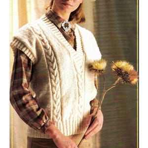 Puede incluir: Un chaleco de punto blanco con cable, cuello en V y una camisa a cuadros marrón y blanca debajo. El chaleco tiene un broche decorativo en el cuello.