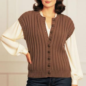 Op de afbeelding: Een mouwloos gebreid vest in bruin met een verticale ribbelstructuur, een ronde halslijn en een knoopsluiting aan de voorzijde met acht bijpassende bruine knopen. Het vest wordt gedragen over een crèmekleurige blouse met lange mouwen en een donkerblauwe jeans.