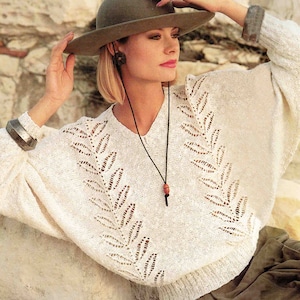 May include: A white knitted jumper with a leaf pattern and a v-neck. The jumper has long sleeves and a relaxed fit. The model is wearing a brown hat and a brown leather necklace.