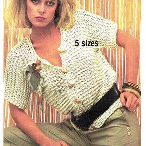 May include: A light beige, open-weave knit top with wooden buttons and a decorative brooch. The top is paired with khaki-colored pants and a black belt. The text "5 sizes" is visible on the top.