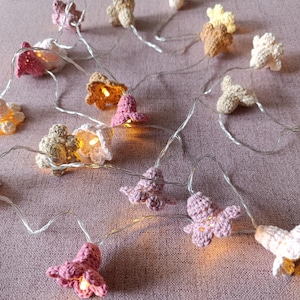 Może przedstawiać: Girlanda z szydełkowych kwiatów w odcieniach różu, beżu i kremu. Każdy kwiat ma małą, ciepłą białą diodę LED. Światła są nawleczone na cienki, srebrny drut, tworząc miękki, fantazyjny blask.