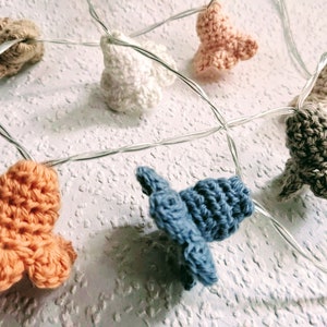 Puede incluir: Una cadena de luces blancas con cubiertas de ganchillo en forma de campana en varios colores, incluyendo beige, melocotón, blanco, gris, azul y naranja.