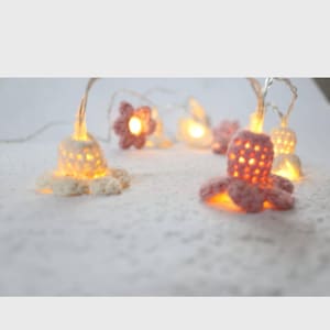 Könnte beinhalten: Eine Lichterkette mit gehäkelten Oktopus- und Blumenformen. Die Oktopus-Lichter sind rosa und weiß, und die Blumenlichter sind rosa. Die Lichter leuchten warmgelb und erzeugen einen skurrilen und dekorativen Effekt.