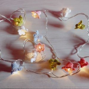 Könnte beinhalten: Eine Lichterkette mit gehäkelten Blumenhüllen in verschiedenen Farben, darunter Rosa, Blau, Grün und Weiß. Die Lichter sind auf einem silbernen Draht aufgefädelt und leuchten, wodurch ein warmes Licht entsteht.