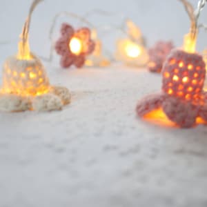 Könnte beinhalten: Eine Lichterkette mit weißen und rosafarbenen gehäkelten Blumen und warmweißen LED-Lampen. Die Lichter sind auf einem durchsichtigen Draht aufgefädelt und leuchten, wodurch ein warmes Licht entsteht.