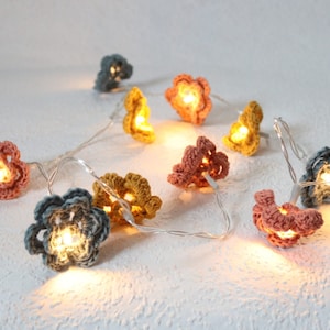 Pode incluir: Uma corda de luzes com capas de flores de crochê em tons de azul, rosa e amarelo. As luzes são enfiadas em um fio transparente e emitem um brilho quente.