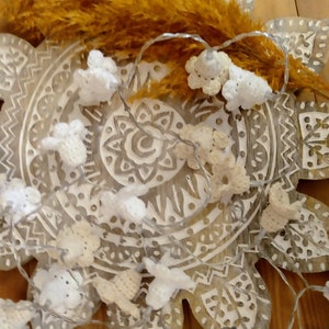 Op de afbeelding: Een string met witte gehaakte bloemenlampjes gewikkeld rond een decoratief houten mandala. De lampjes zijn aan een doorzichtig draad geregen en hebben kleine LED-lampjes.