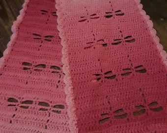 May include: A pink crocheted scarf with a dragonfly pattern. The scarf is made with a gradient yarn, starting with a lighter pink and fading to a darker pink.