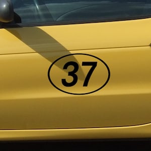 Könnte beinhalten: Ein gelbes Auto mit einem schwarzen ovalen Aufkleber, auf dem "37" steht, an der Seite.