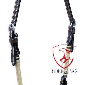 Puede incluir: Cabestro de cuero marrón con una cabezada de cuerda trenzada blanca. El cabestro tiene una hebilla plateada y está cosido con hilo blanco. El cabestro está etiquetado como "Rider Vivan".