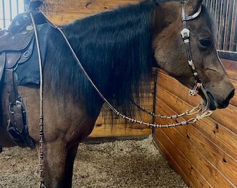 Könnte beinhalten: Ein braunes Pferd mit schwarzer Mähne und Schweif trägt einen braunen Ledersattel und ein Zaumzeug mit einer perlenbesetzten Zügel. Das Pferd steht in einem Stall mit Holzwänden.