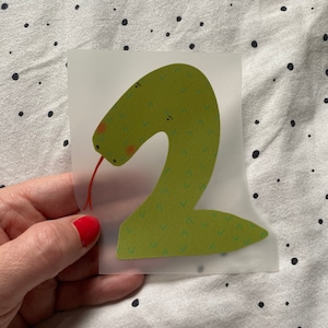 Könnte beinhalten: Eine grüne Schlange in Form der Zahl Zwei mit einer roten Zunge. Die Schlange hat ein Muster aus kleinen grünen und blauen Formen.