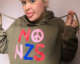 May include: A brown hooded sweatshirt with the words "NO NZS" printed on the front in pink, red, and blue paint. A pink peace sign is between the words "NO" and "NZS".