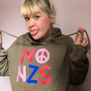 Puede incluir: Una sudadera con capucha marrón con las palabras "NO NZS" impresas en la parte delantera en pintura rosa, roja y azul. Un símbolo de paz rosa se encuentra entre las palabras "NO" y "NZS".
