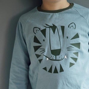 May include: A light blue long-sleeved shirt with a green tiger face graphic. The shirt has a green trim around the neck and cuffs.
