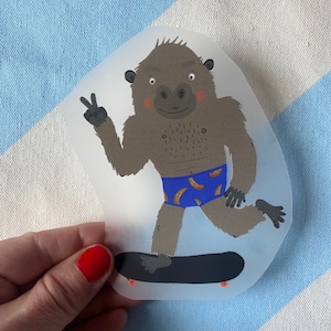 Könnte beinhalten: Ein brauner Gorilla, der blaue und braune Badehosen trägt, fährt auf einem schwarzen Skateboard mit orangefarbenen Rädern. Der Gorilla zeigt mit einer Hand ein Peace-Zeichen und hat einen fröhlichen Gesichtsausdruck.