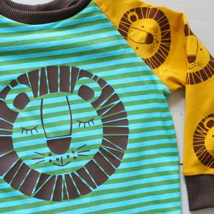 May include: A blue and green striped long-sleeved shirt with a brown lion face on the front and brown lion faces on the yellow sleeves.