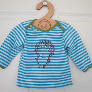 May include: A baby's long-sleeved top with blue and white horizontal stripes. The shirt has a cartoon hedgehog graphic on the front. The neckline and cuffs are trimmed with green fabric. It hangs on a wooden hanger.