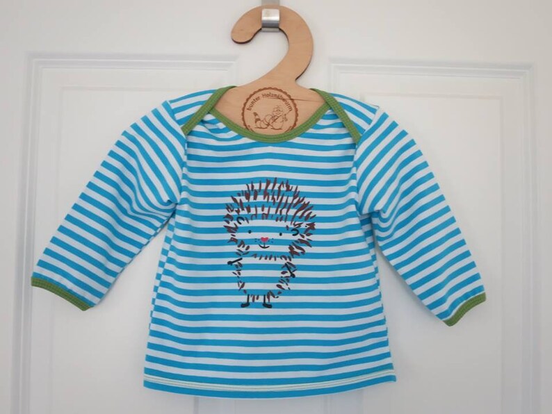 May include: A baby's long-sleeved top with blue and white horizontal stripes. The shirt has a cartoon hedgehog graphic on the front. The neckline and cuffs are trimmed with green fabric. It hangs on a wooden hanger.