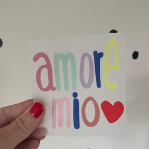 Peut inclure: Un autocollant carré avec les mots "amore mio" en lettres colorées. Le mot "amore" est en rose, vert menthe, violet et jaune. Le mot "mio" est en rose, bleu et marron, avec un cœur rouge.