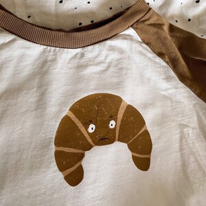 Könnte beinhalten: Ein weißes T-Shirt mit braunen Ärmeln und einem braunen Comic-Croissant mit einem Gesicht auf der Vorderseite.