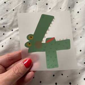 Pode incluir: Adesivo verde em forma de crocodilo com o número quatro. O crocodilo tem a boca vermelha e os olhos amarelos.