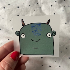 Könnte beinhalten: Ein grüner Monster-Aufkleber mit blauen und braunen Details. Das Monster hat zwei Hörner, große Augen und einen lächelnden Mund.