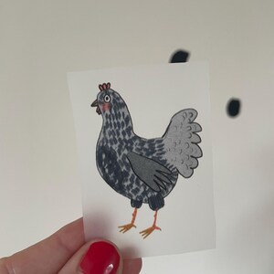 Può includere: Un piccolo adesivo rettangolare con un'illustrazione di un pollo dei cartoni animati. Il pollo è principalmente grigio con macchie nere, una cresta rossa e zampe arancioni. L'adesivo è tenuto su uno sfondo bianco con pois neri.